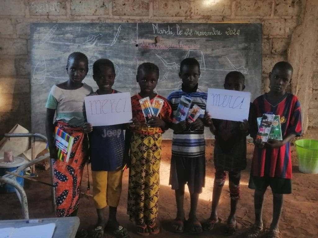 A group of six young students in Dar Dalam, Kedougou, Senegal, showcase new school supplies made possible through a Small Change Better World grant. Two hold signs saying "Merci"
